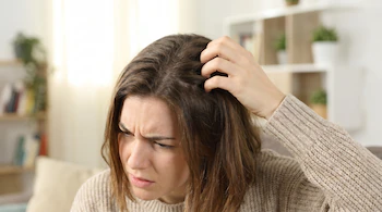 Una mujer con cabello castaño y suéter beige se rasca el cuero cabelludo con la mano derecha, mientras frunce el ceño.