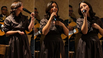 Ángela Aguilar con una blusa y falda marrones, canta en un micrófono. Detrás, dos músicos de mariachi tocan guitarras