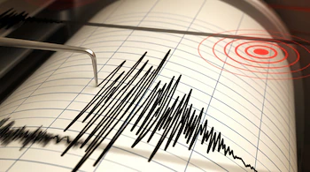 Un sismógrafo registra ondas sísmicas en un papel cuadriculado blanco. Se observa un brazo metálico con una punta que dibuja líneas negras, y círculos rojos concéntricos.