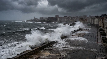 Olas rompen contra un malecón e inundan una calle urbana bajo un cielo gris con lluvia